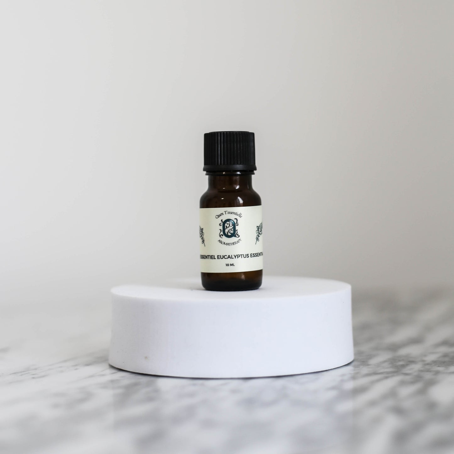 10ml amber bottle of Eucalyptus Essential Oil by Quen T’essentielle, displayed on a minimalist white pedestal with clean marble background – natural remedy for respiratory support and mental clarity.
