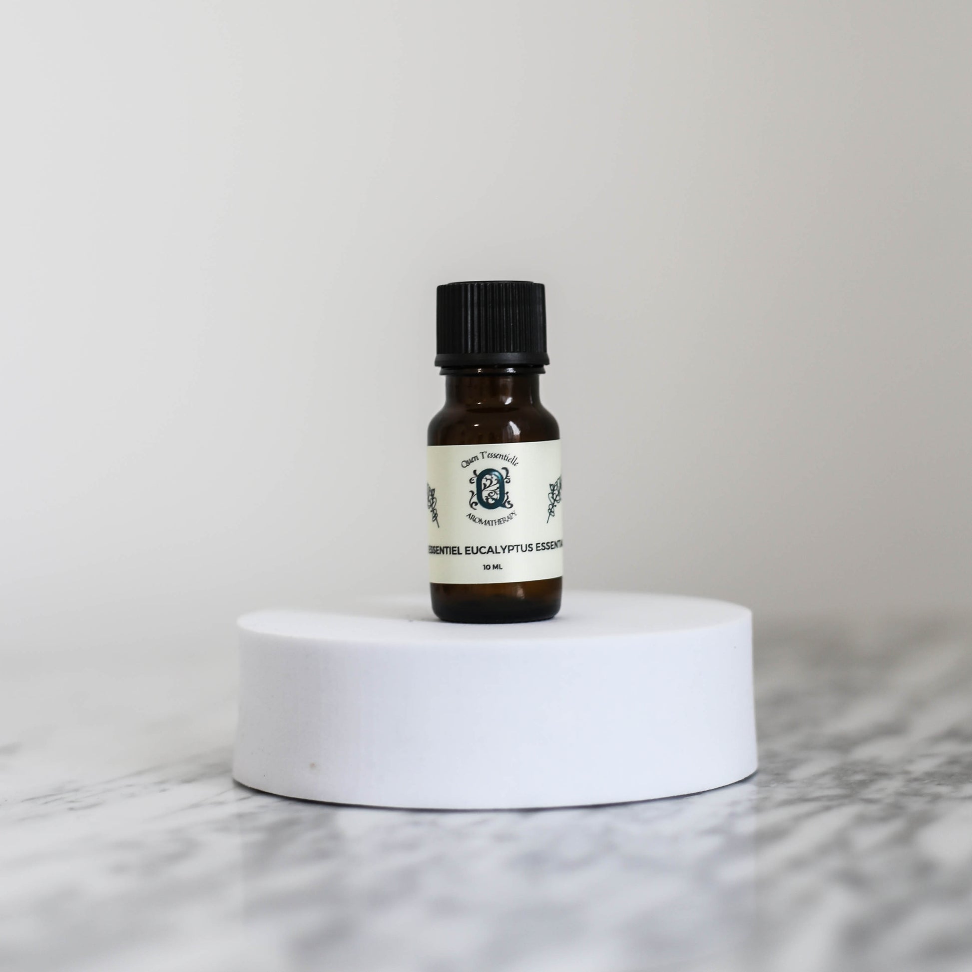 10ml amber bottle of Eucalyptus Essential Oil by Quen T’essentielle, displayed on a minimalist white pedestal with clean marble background – natural remedy for respiratory support and mental clarity.