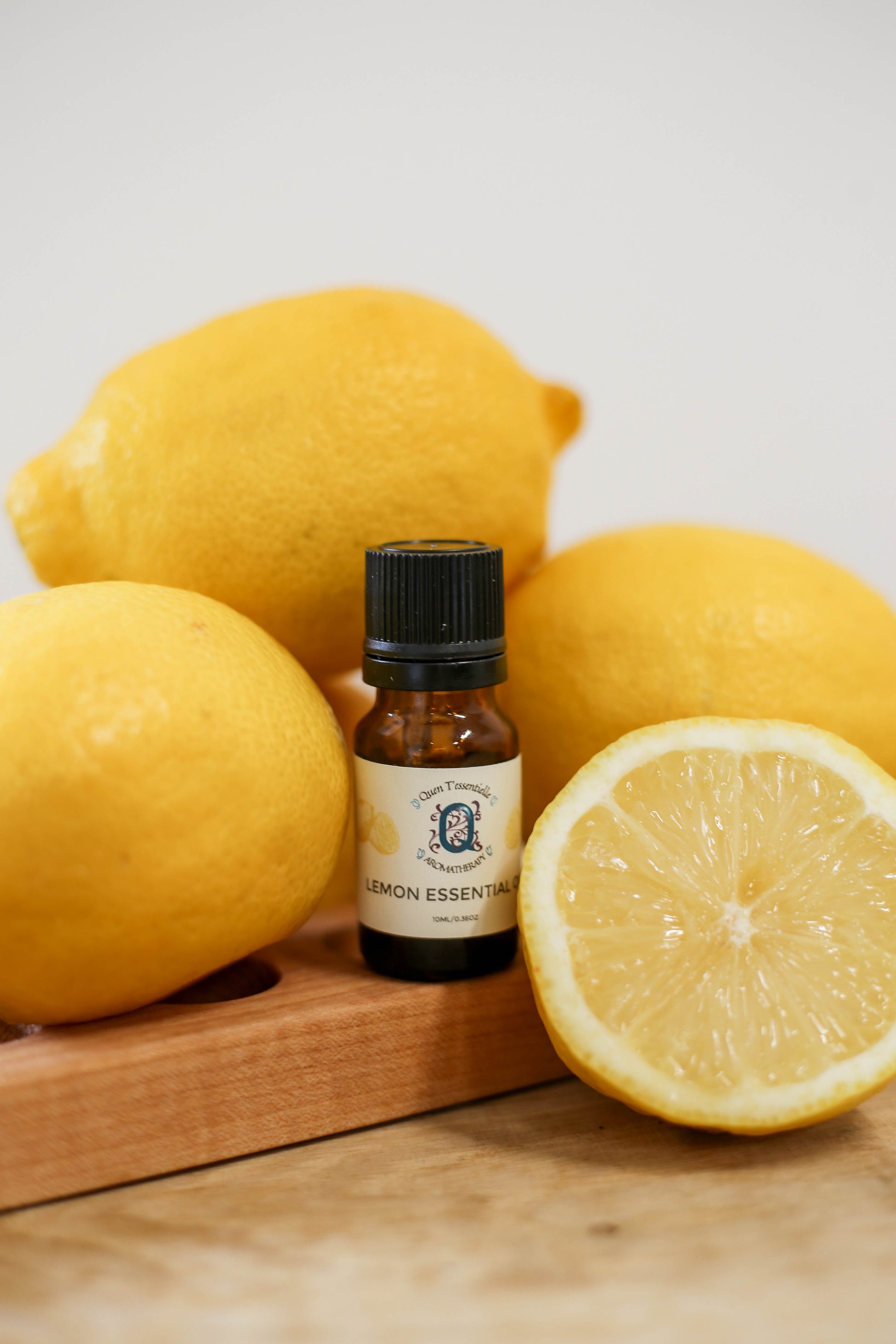 A 15 ml amber bottle of Lemon Essential Oil by Quen T’essentielle Aromatherapy, surrounded by whole and halved fresh lemons on a wooden surface, capturing a vibrant and zesty citrus aesthetic.