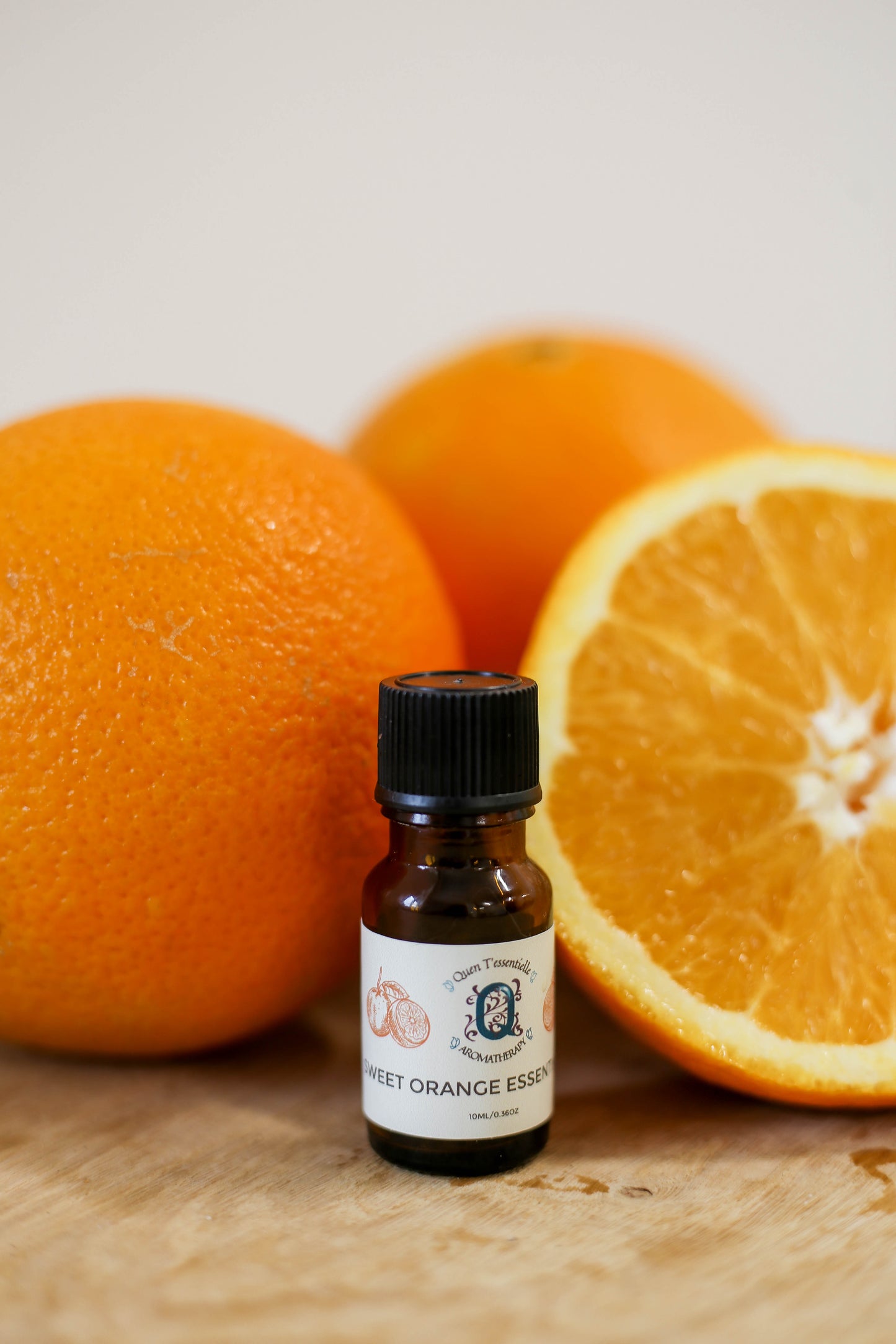 A 10 ml amber bottle of Sweet Orange Essential Oil by Quen T’essentielle Aromatherapy, displayed in front of fresh whole and halved oranges on a wooden surface, evoking a bright and citrusy vibe.
