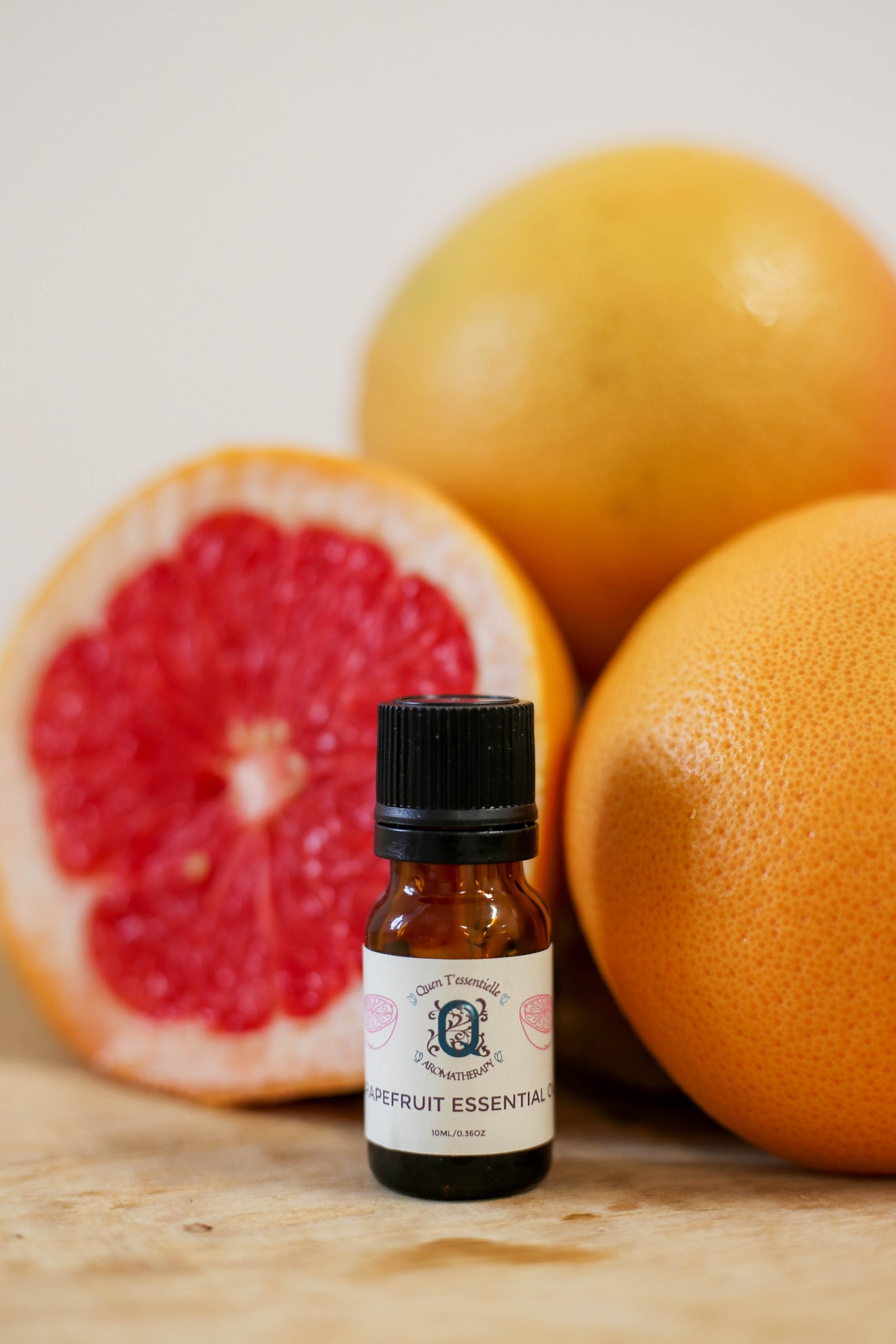 A 10 ml amber bottle of Grapefruit Essential Oil by Quen T’essentielle Aromatherapy, displayed in front of fresh whole and halved grapefruits on a wooden surface, evoking a bright and citrusy vibe.