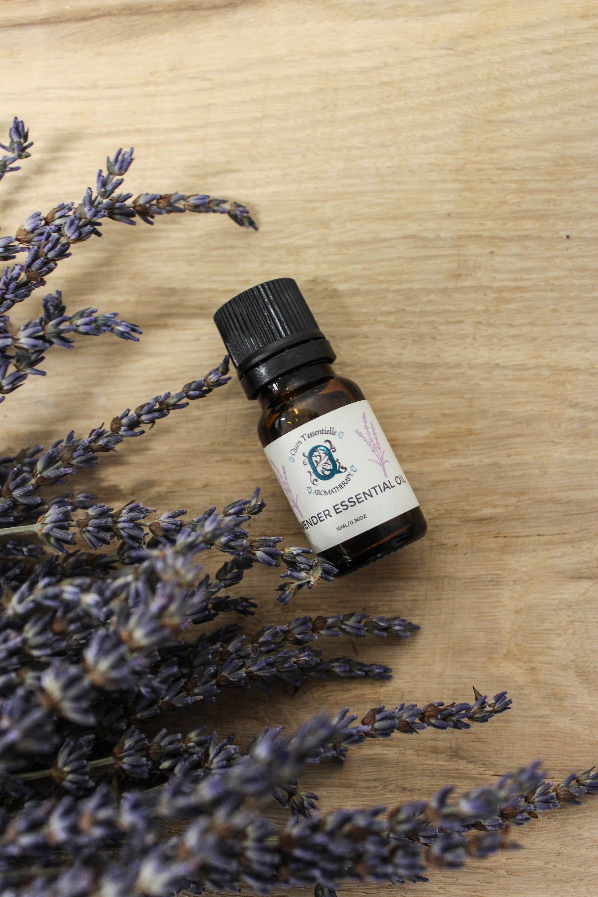 A 10 ml amber glass bottle of Lavender Essential Oil by Quen T’essentielle Aromatherapy, displayed on a wooden surface surrounded by dried lavender sprigs, creating a calm and botanical aesthetic.