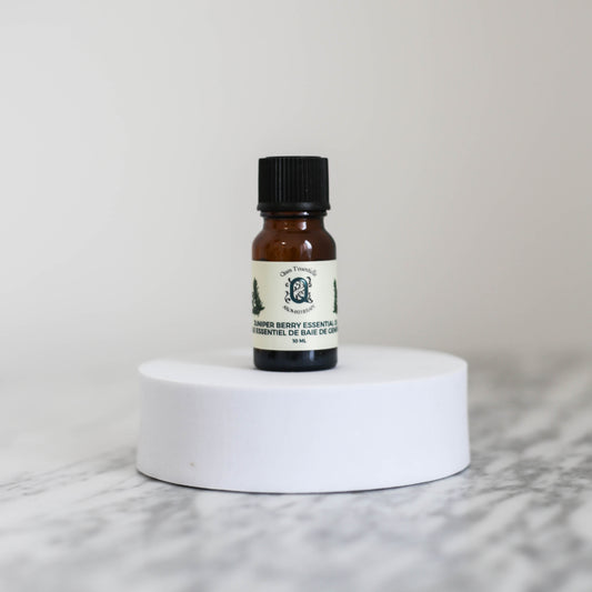 A 10 ml amber glass bottle of Juniper Berry Essential Oil by Quen T’essentielle Aromatherapy, displayed on a white circular pedestal with a minimalist marble background.