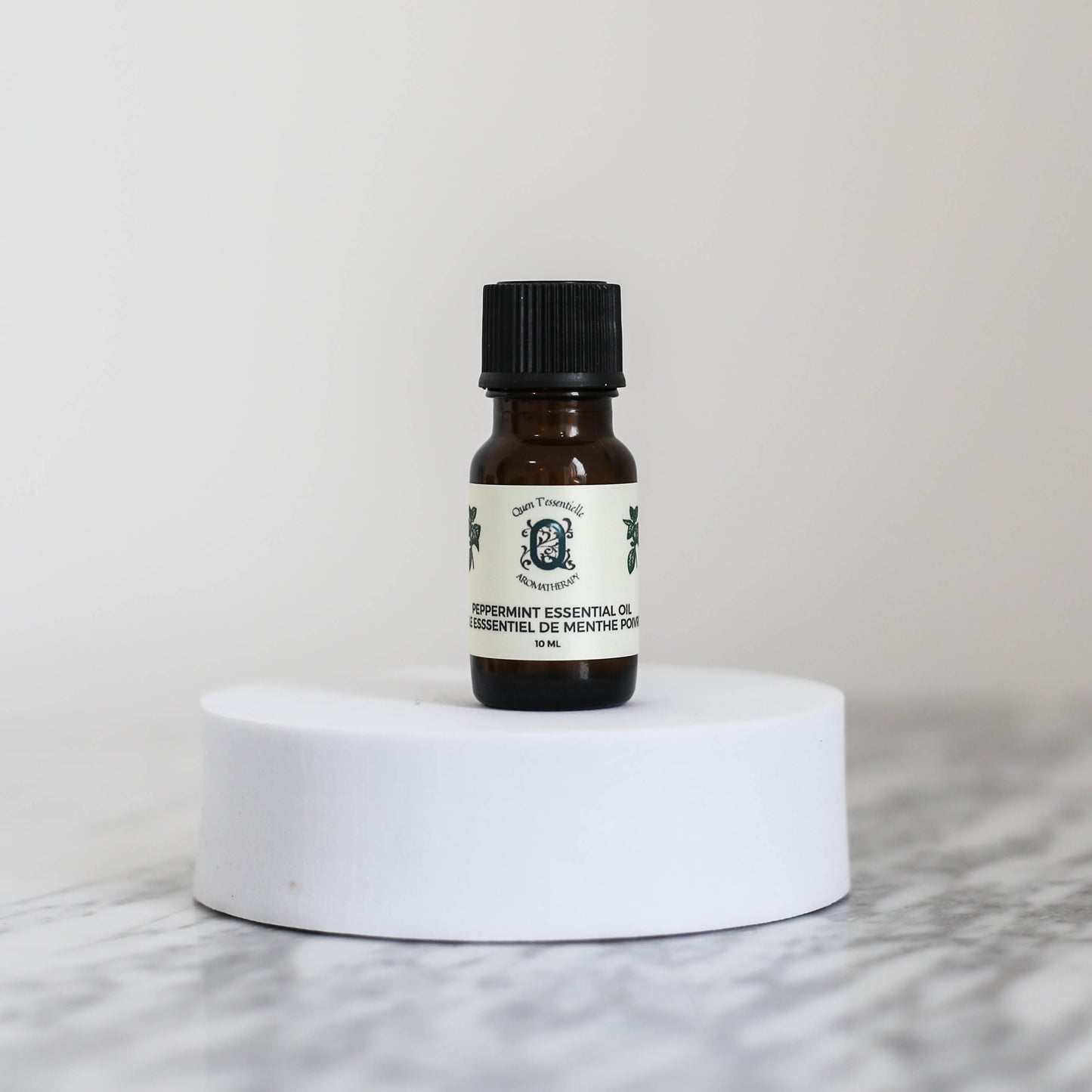 A 10 ml amber glass bottle of Peppermint Essential Oil by Quen T’essentielle Aromatherapy, displayed on a white circular pedestal with a minimalist marble background