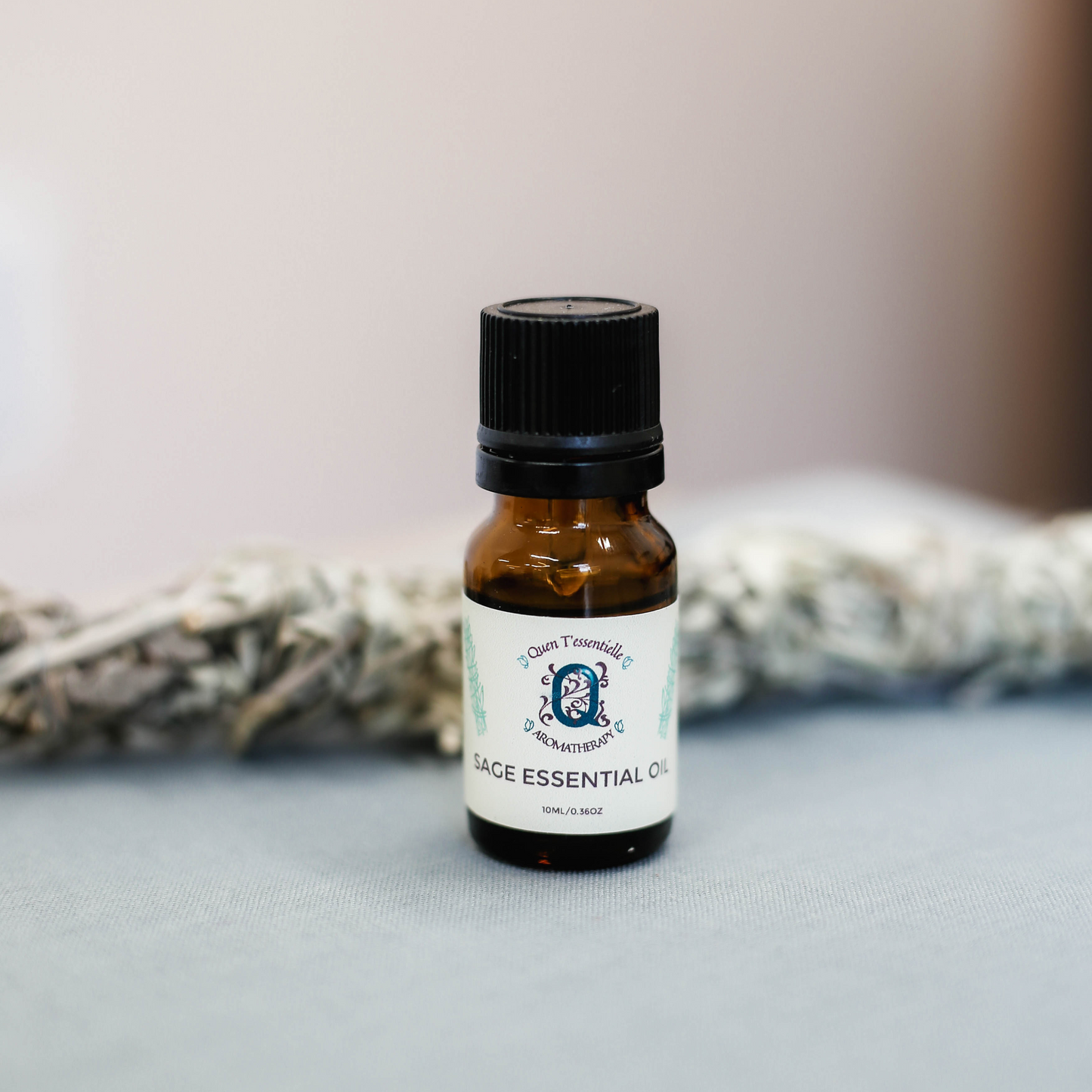 A 10 ml amber glass bottle of Sage Essential Oil by Quen T’essentielle Aromatherapy, standing in front of a sage bundle on a soft grey surface, evoking a calming and purifying atmosphere.