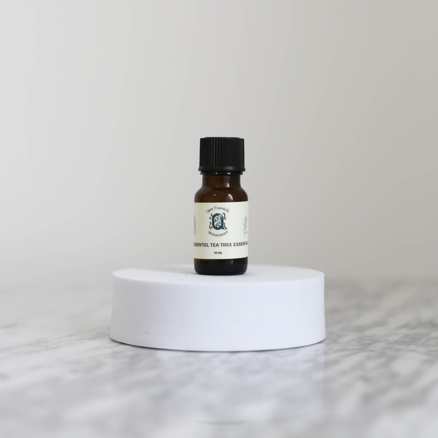 A 10 ml amber glass bottle of Tea Tree Essential Oil by Quen T’essentielle Aromatherapy, displayed on a white circular pedestal with a minimalist marble background.