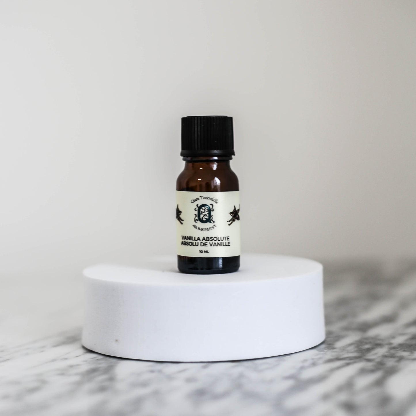 A 10 ml amber glass bottle of Vanilla Absolute Oil by Quen T’essentielle Aromatherapy, displayed on a white circular pedestal with a minimalist marble background.