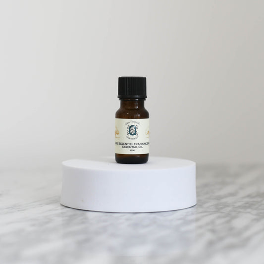 A 10 ml amber glass bottle of Frankincense Essential Oil by Quen T’essentielle Aromatherapy, displayed on a white circular pedestal with a minimalist marble background.