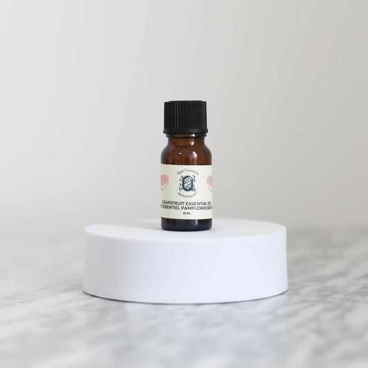 A 10 ml amber glass bottle of Grapefuit Essential Oil by Quen T’essentielle Aromatherapy, displayed on a white circular pedestal with a minimalist marble background.