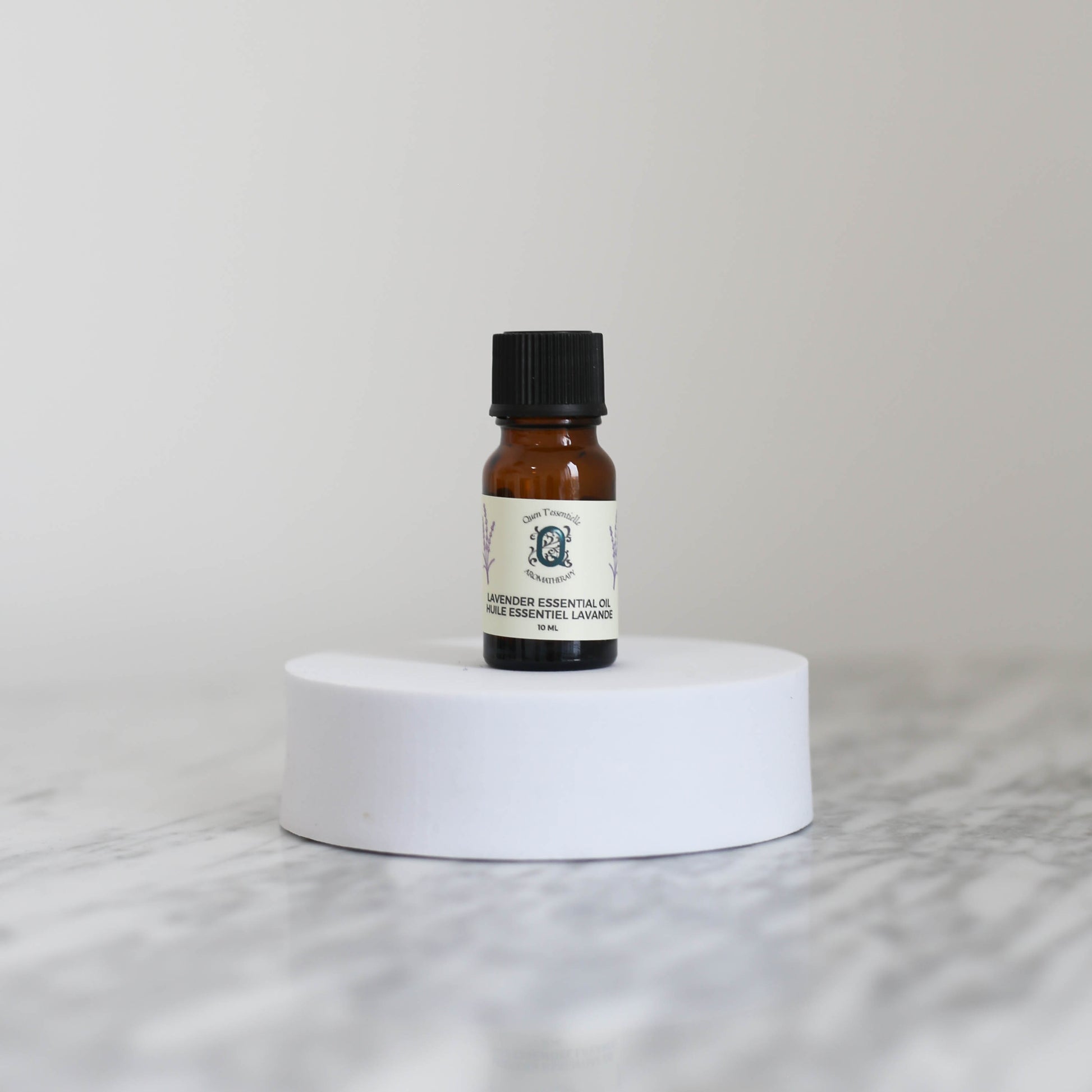 A 10 ml amber glass bottle of Lavender Essential Oil by Quen T’essentielle Aromatherapy, displayed on a white circular pedestal with a minimalist marble background.