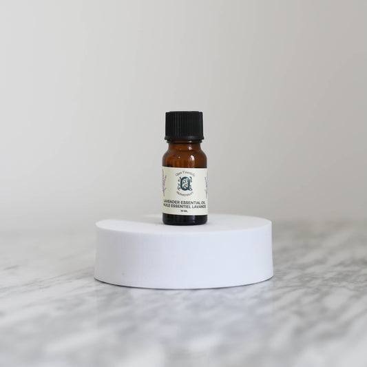 A 10 ml amber glass bottle of Lavender Essential Oil by Quen T’essentielle Aromatherapy, displayed on a white circular pedestal with a minimalist marble background.