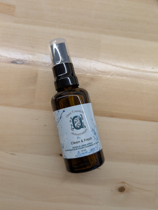 A brown glass spray bottle labeled "Quen T'essentielle Aromatherapy" is displayed on a light wooden surface. The label reads "Clean & Fresh" and describes the product as a "Room & Linen Spray" in both English and French ("Vaporisateur pour chambre et draps"). The bottle contains 50 mL of product and features decorative blue water splash graphics around the label. The spray nozzle is black with a clear plastic cap.