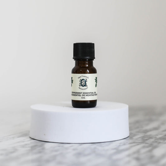 A 10 ml amber glass bottle of Peppermint Essential Oil by Quen T’essentielle Aromatherapy, displayed on a white circular pedestal with a minimalist marble background