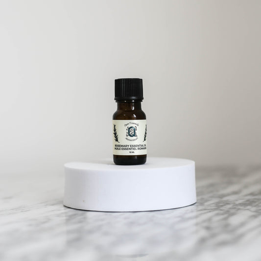 A 10 ml amber glass bottle of Rosemary Essential Oil by Quen T’essentielle Aromatherapy, displayed on a white circular pedestal with a minimalist marble background