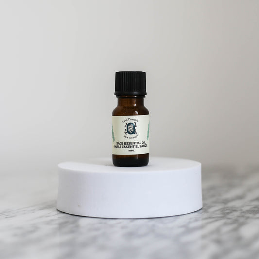 A 10 ml amber glass bottle of Sage Essential Oil by Quen T’essentielle Aromatherapy, displayed on a white circular pedestal with a minimalist marble background
