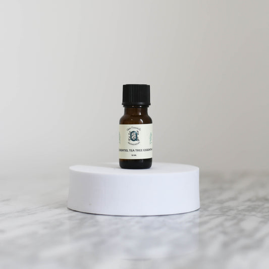 A 10 ml amber glass bottle of Tea Tree Essential Oil by Quen T’essentielle Aromatherapy, displayed on a white circular pedestal with a minimalist marble background.