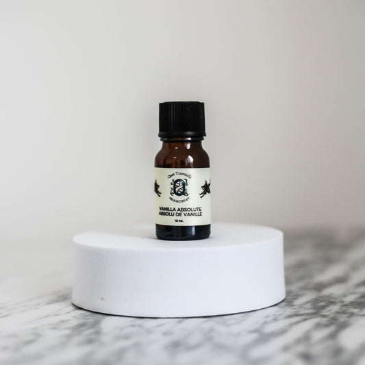A 10 ml amber glass bottle of Vanilla Absolute Oil by Quen T’essentielle Aromatherapy, displayed on a white circular pedestal with a minimalist marble background.