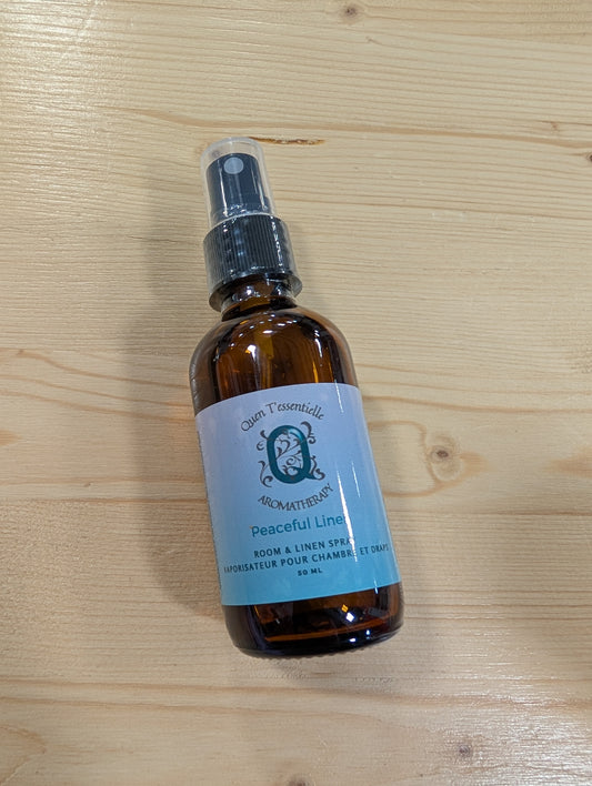 A brown glass spray bottle is shown resting on a light wooden surface. The label on the bottle reads "Quen T'essentielle Aromatherapy" with the scent name "Peaceful Linen" printed beneath. It is identified as a "Room & Linen Spray" and includes the French translation "Vaporisateur pour chambre et draps." The bottle contains 50 mL of product and features a black spray nozzle with a clear plastic cap. The label design is soft blue with an elegant, calming aesthetic.