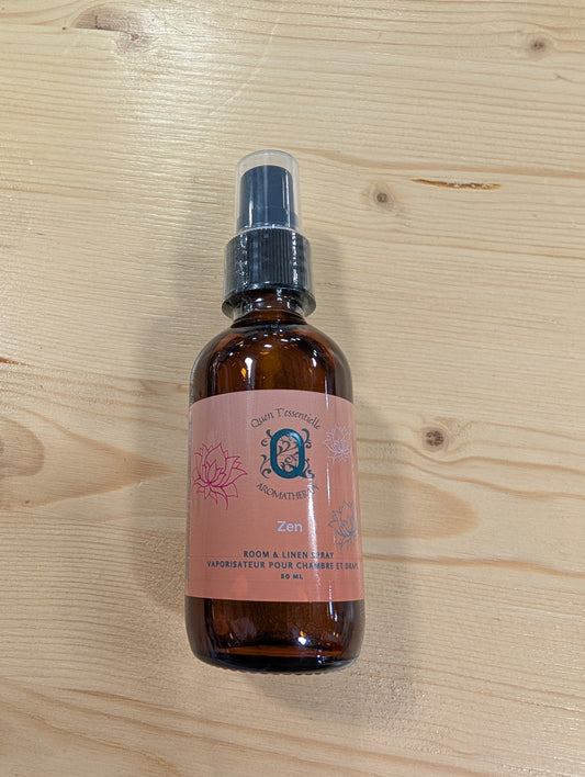 A brown glass spray bottle is placed on a light wooden surface. The label, which is an orange color that lightens up to a pink towards the top, reads 'Quen T’essentielle Aromatherapy' and features the scent name 'Zen.' It is described as a 'Room & Linen Spray' with the French translation 'Vaporisateur pour chambre et draps.' The bottle contains 50 mL of product. There is a dark pink lotus outline on the left-hand side, a smaller, light pink one on the right, and a teal lotus flower below the light pink one,