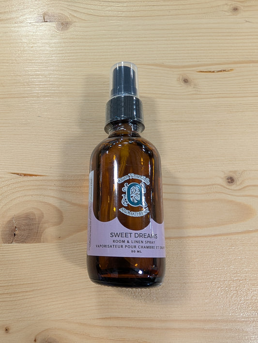 A brown glass spray bottle is placed on a light wooden surface. The label reads "Quen T’essentielle Aromatherapy" and features the scent name "Sweet Dreams." It is described as a "Room & Linen Spray" with the French translation "Vaporisateur pour chambre et draps." The bottle contains 50 mL of product. The label is a soft pink color, complementing the calming theme suggested by the scent name. The spray nozzle is black with a clear plastic cap.